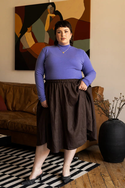 Woman wearing the Tuesday Skirt by Lights of All in Cocoa with the Essential turtleneck in Violet standing in a living room with abstract art on the wall.
