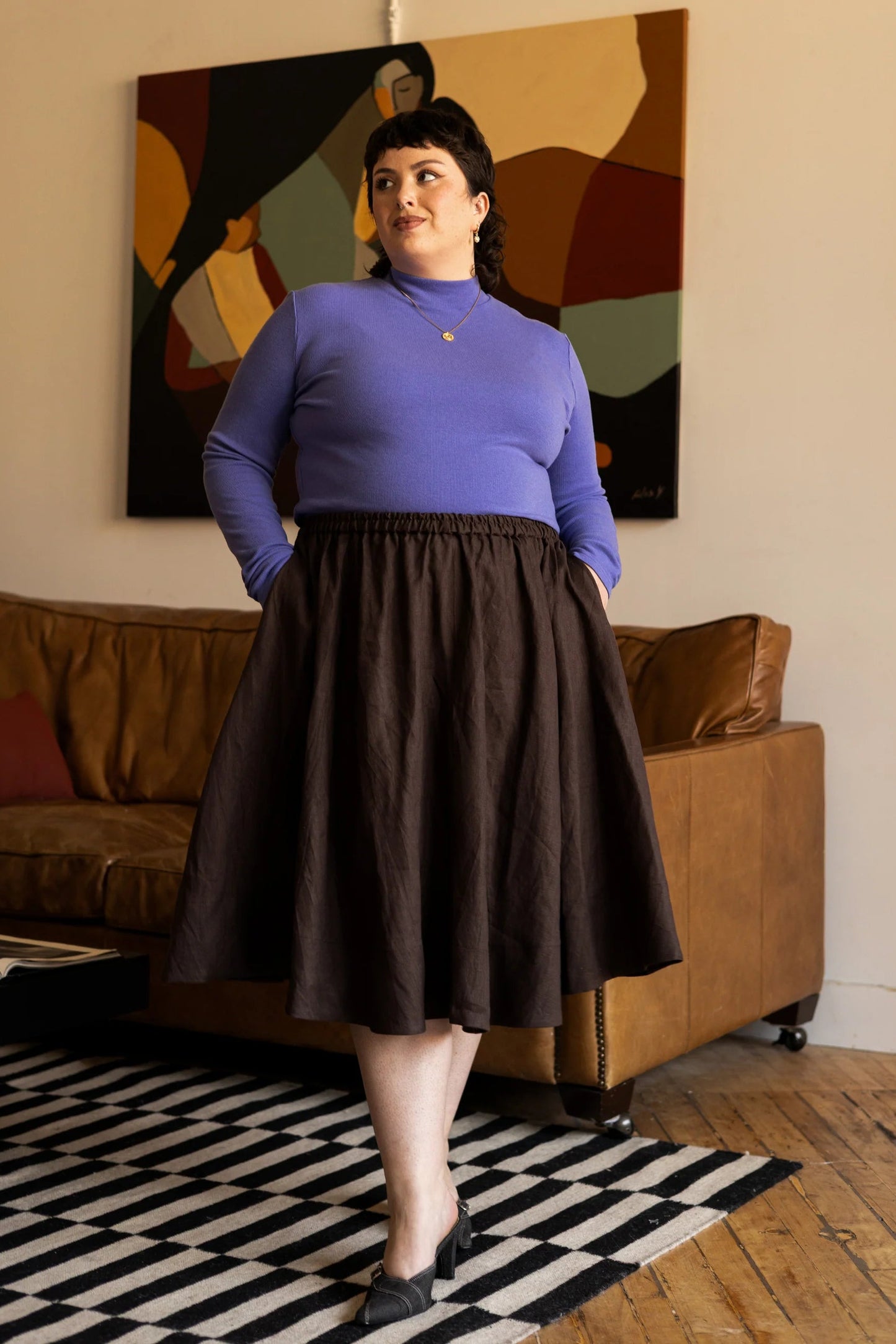 Woman wearing the Tuesday Skirt by Lights of All in Cocoa with the Essential turtleneck in Violet standing in a living room with abstract art on the wall.