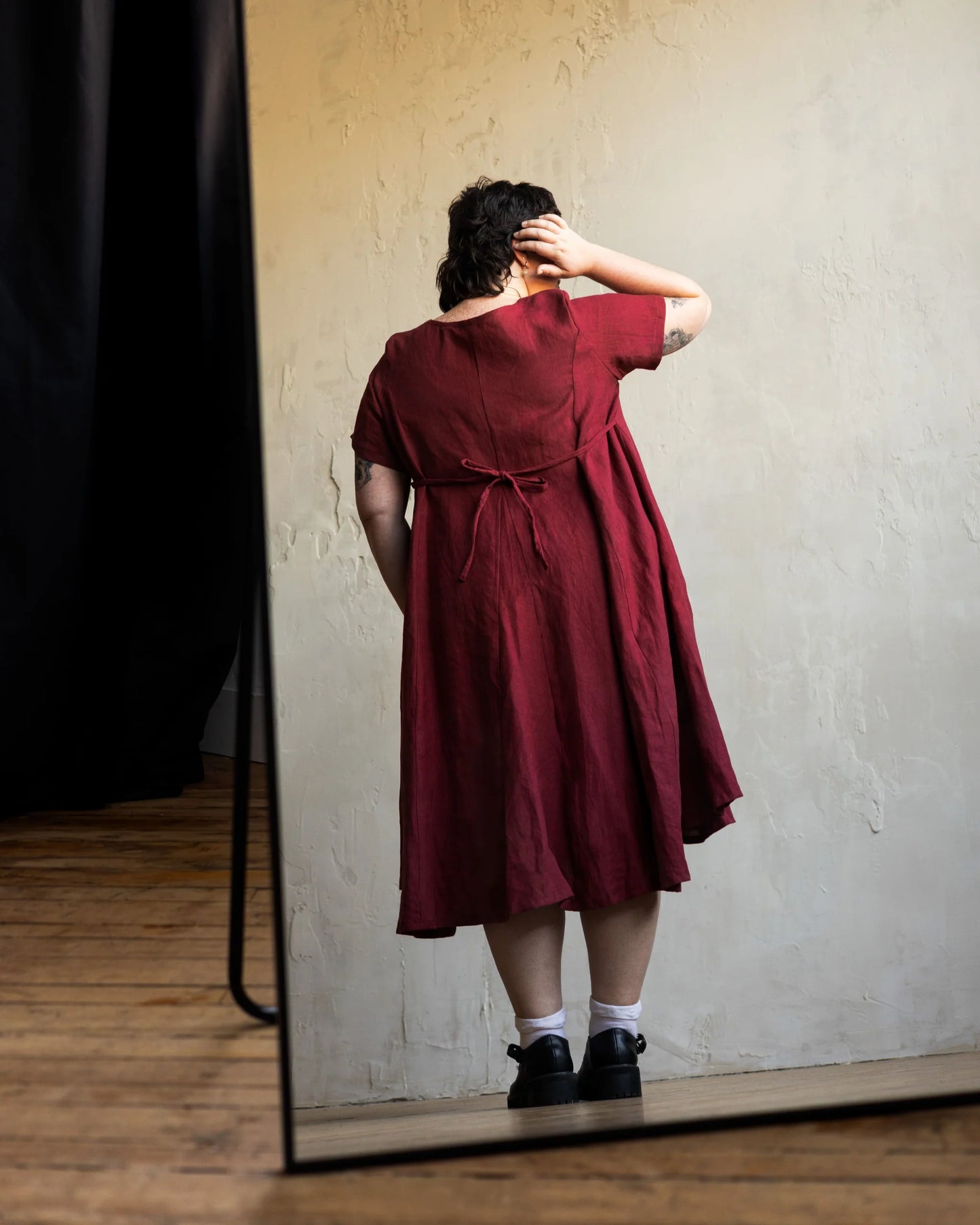 Back view of a woman wearing the Astoria Dress by Lights of All in Garnet, shown in a mirror 