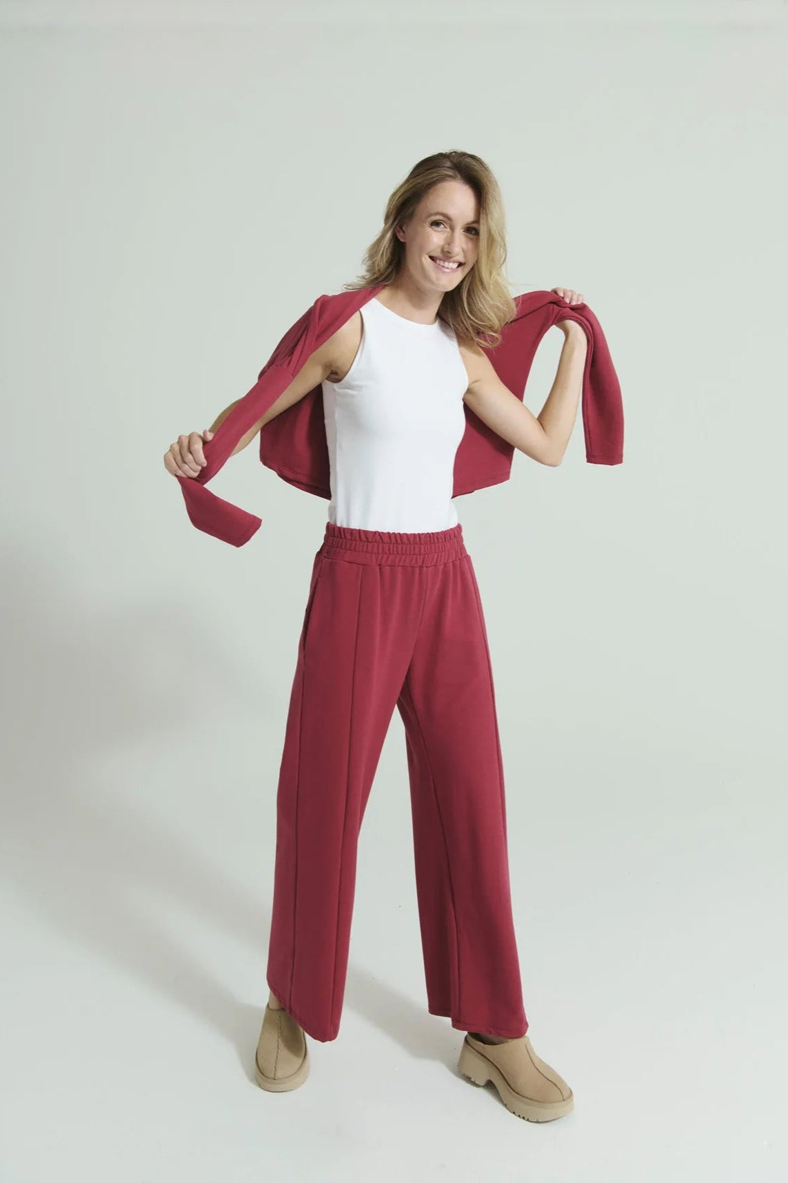 A woman wearing the Romaine Sweater and Tadoussac Pants by Message Factory in Raspberry: a sweatshirt with a funnel neck and long drop sleeves and flared sweatpants with an elastic waist, both in a bamboo/cotton blend. She is wearing a white tank with them and is standing in front of a white background. 