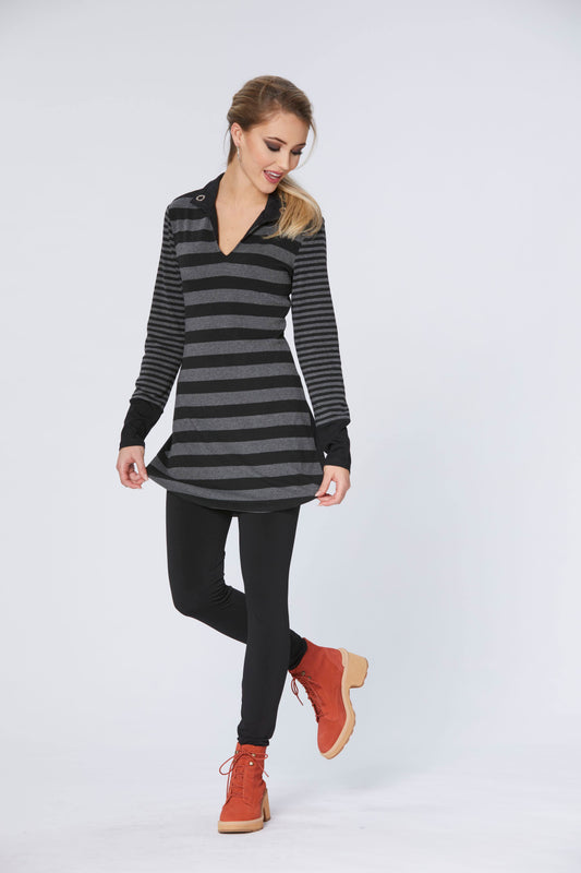 A woman wearing the Swift Tunic by Luc Fontaine, with a v-neck with a collar, wide black and grey stripes on the body, narrow black and grey stripes on the sleeves, and contrasting black cuffs, with black leggings. She is standing in front of a white background 