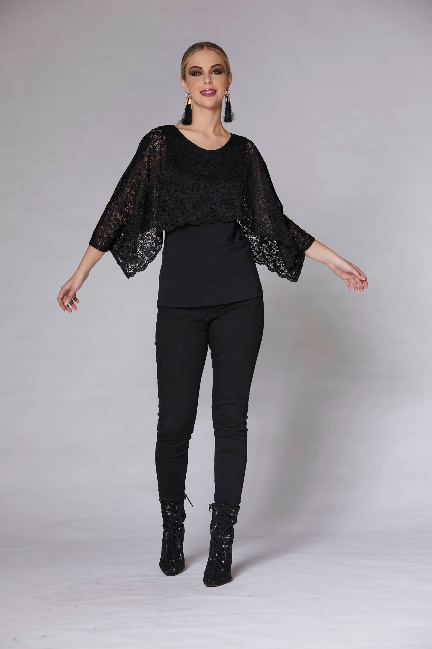 A woman wearing the Chriss Top by Luc Fontaine in Black, with a solid black tank and a lace overlay that falls over the bust and down to the lower arms, with black pants. She is standing in front of a grey background.