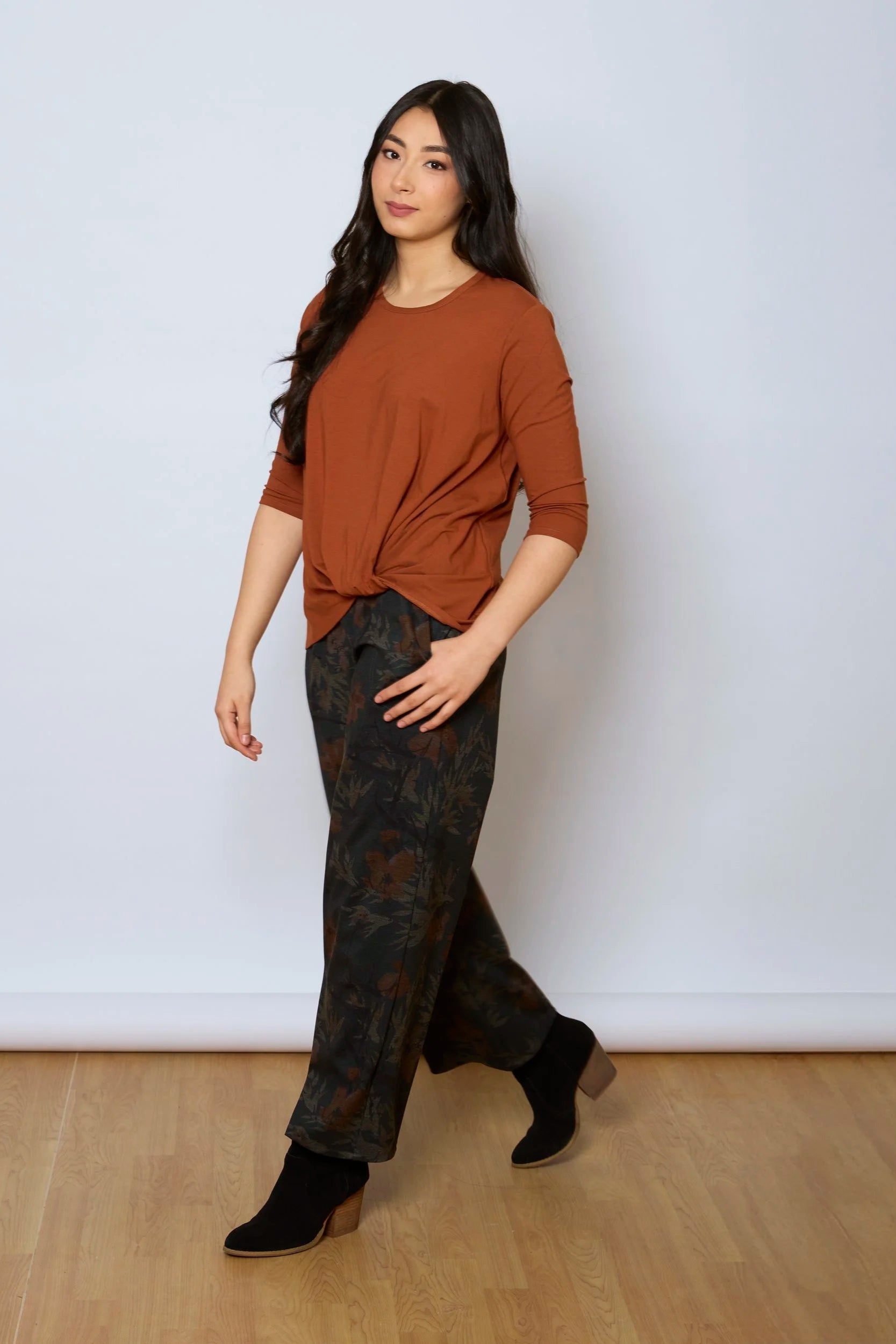 A woman wearing the Luisa Pants by Tangente in Floral, featuring an elastic waist with pleats, straight legs, and side pockets. She is wearing them with a rust coloured top and standing in front of a white background. 