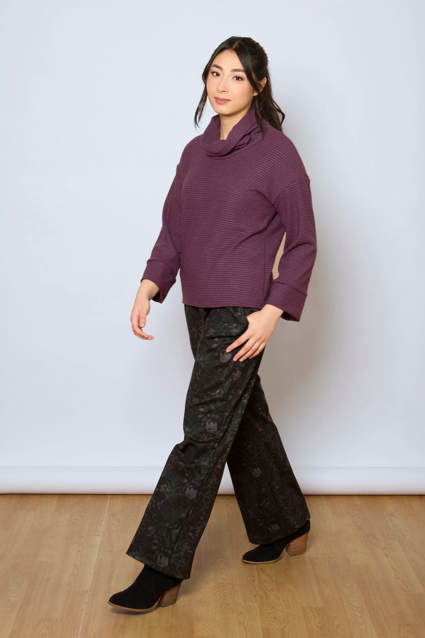 A woman wearing the Luisa Pants by Tangente in Woodland print, featuring an elastic waist with pleats, straight legs, and side pockets. She is wearing them with a purple top and standing in front of a white background. 
