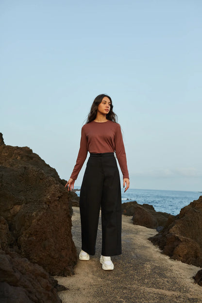 A woman waering the Vienna Pants by MAS in Pepper, wide legged pants with a high waist and a zip front. She is wearing them with a brown top and standing on a rocky path by the water. 