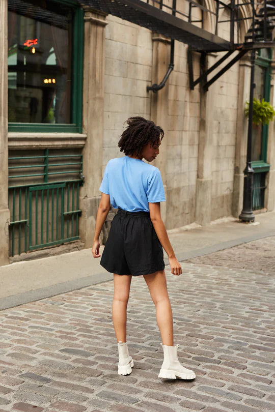 Back view of a woman wearing the Cali Short by MAS in Pepper with a blue top, standing on a cobblestone street