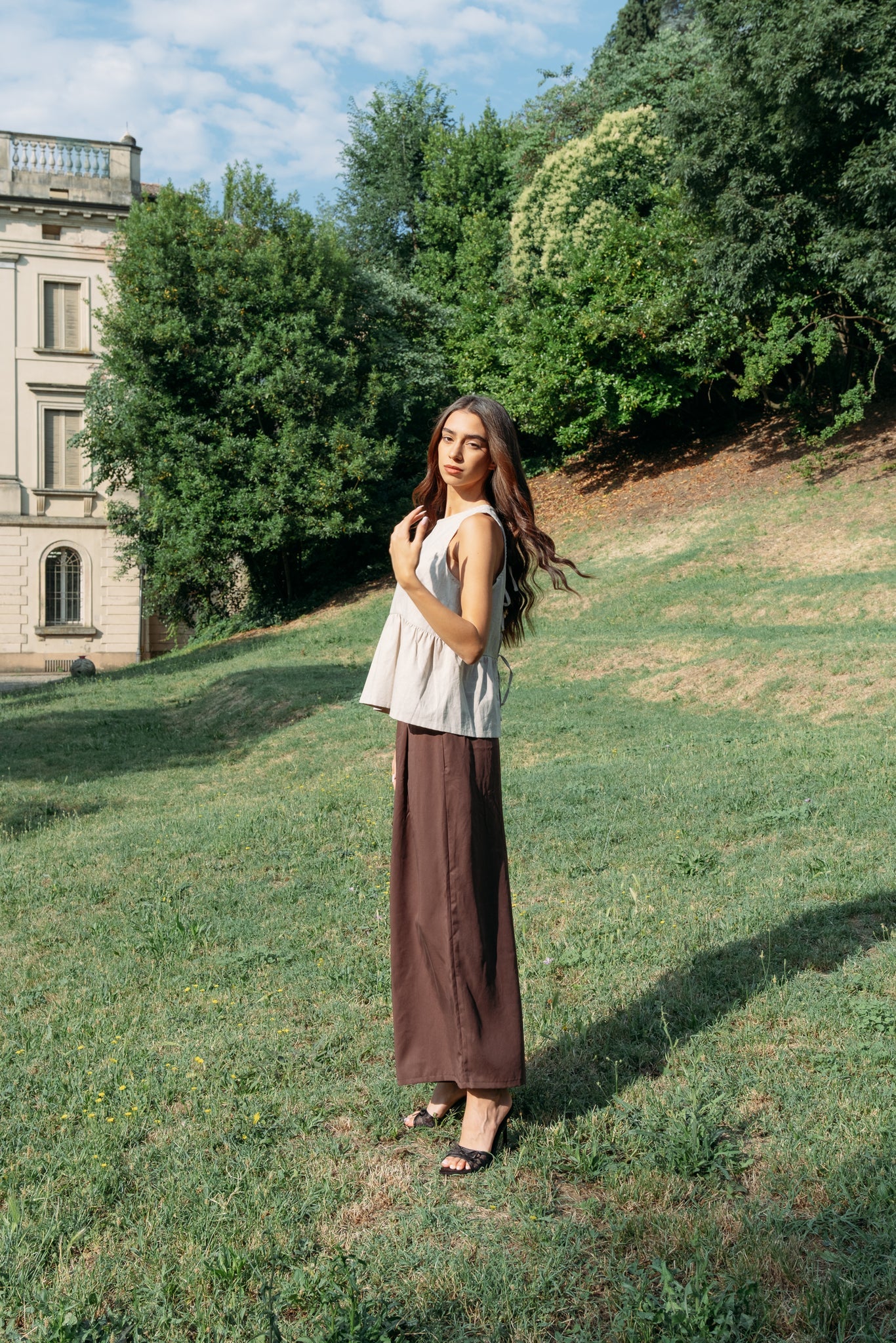 A woman wearing the Siena Pants by MAS in Cocoa, wide legged trousers with an elastic waist. She is wearing them with a white tank and standing outdoors by a historic building.