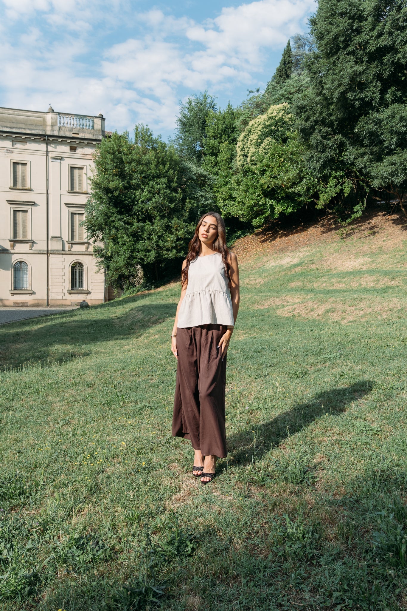 A woman wearing the Siena Pants by MAS in Cocoa, wide legged trousers with an elastic waist. She is wearing them with a white tank and standing outdoors by a historic building.