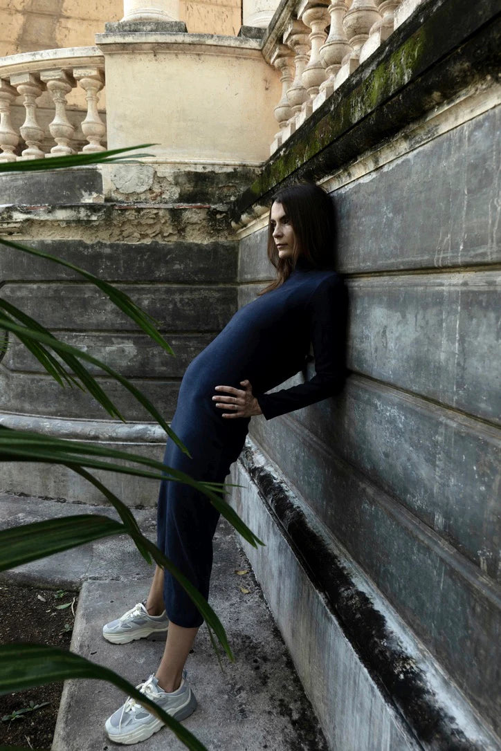 Woman in wearing the Mecca Dress by MAS in Blueberry, a long slim-fitting  turtleneck dress. She is standing next to a stone wall.