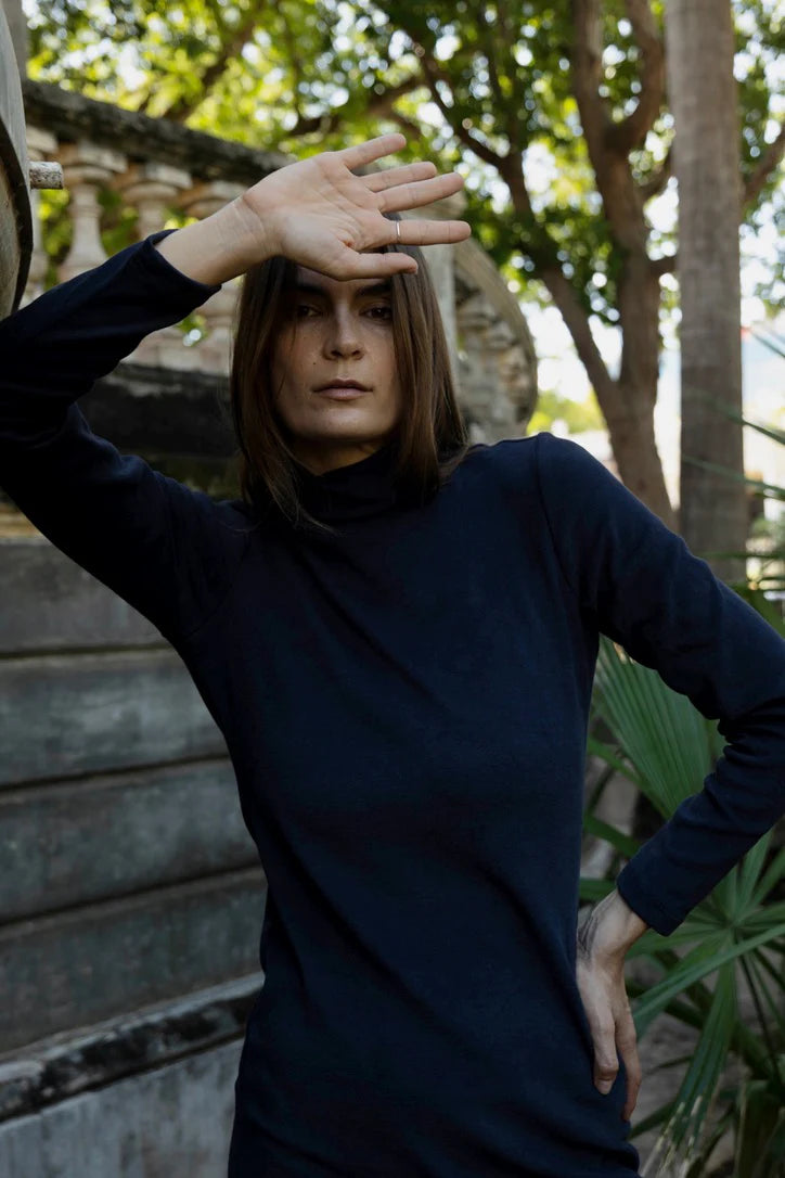 Woman in wearing the Mecca Dress by MAS in Blueberry, a long slim-fitting  turtleneck dress. She is standing next to a stone wall with greenery in the background. 