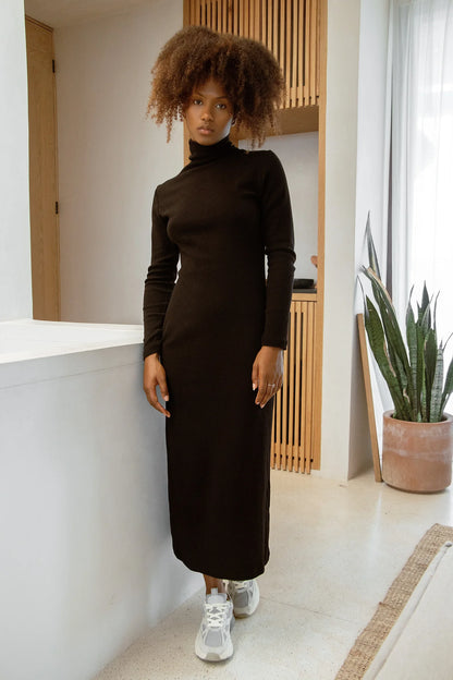 Woman in wearing the Mecca Dress by MAS in Pepper a long slim-fitting  turtleneck dress. She is standing next to a white counter.

