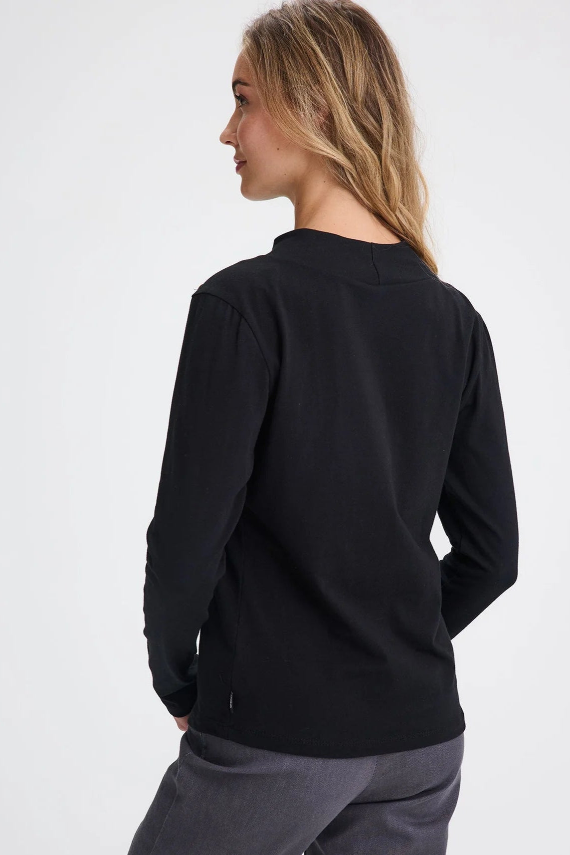 Back view of a woman wearing the Carleton Top by Message Factory in a Black, a long-sleeve top with button details on the shoulder. She is wearing it with grey bottoms and standing in front of a white background. 
