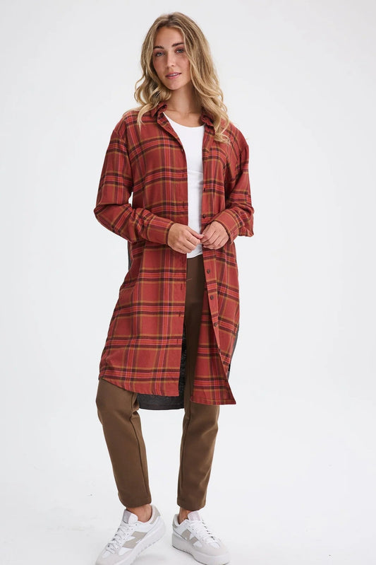 A woman wearing the Donnacona Shirt Dress by Message Factory in Rust Check, a knee-length dress with a button front and collar. She is wearing it open over a white t-shirt and brown pants and standing in front of a white background. 