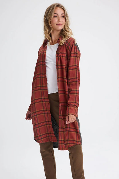 A woman wearing the Donnacona Shirt Dress by Message Factory in Rust Check, a knee-length dress with a button front and collar. She is wearing it open over a white t-shirt and brown pants and standing in front of a white background. 