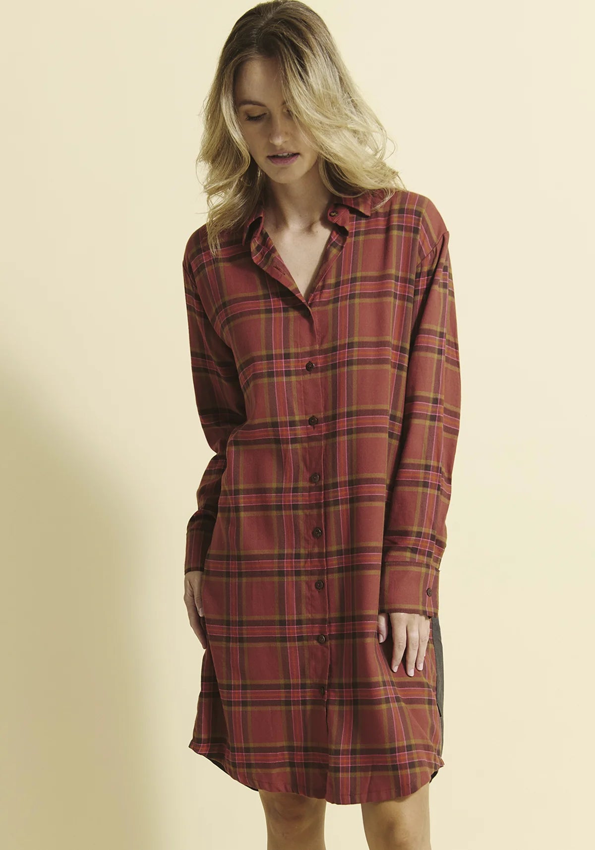 A woman wearing the Donnacona Shirt Dress by Message Factory in Rust Check, a knee-length dress with a button front and collar, standing in front of a yellow background. 