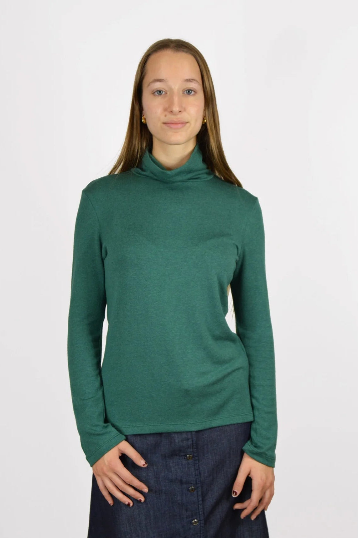 A woman wearing the Gatineau Turtleneck by Message Factory in Green, a classic turtleneck in a bamboo and cotton blend, with a denim skirt. She's standing in front of a white background. 
