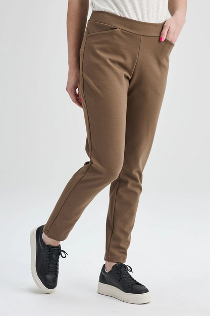 Waist down view of a woman wearing the Sherbrooke Pants by Message Factory in Caramel, pull-on stretch pants with a slim fit, smooth waistband, and two pockets. She is wearing them with a white top and standing in front of a white background. 