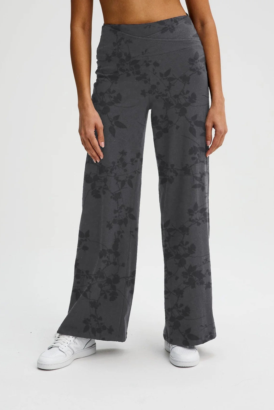 Waist down view of a woman wearing the Villeray Palazzo Pants by Message Factory in Grey Floral Print, wide legged organic cotton pants with a pull-on elastic waistband with a cross-over detail. She is standing in front of a white background. 