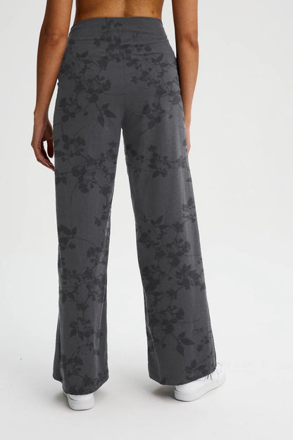 Back waist down view of a woman wearing the Villeray Palazzo Pants by Message Factory in Grey Floral Print, wide legged organic cotton pants with a pull-on elastic waistband with a cross-over detail. She is standing in front of a white background. 