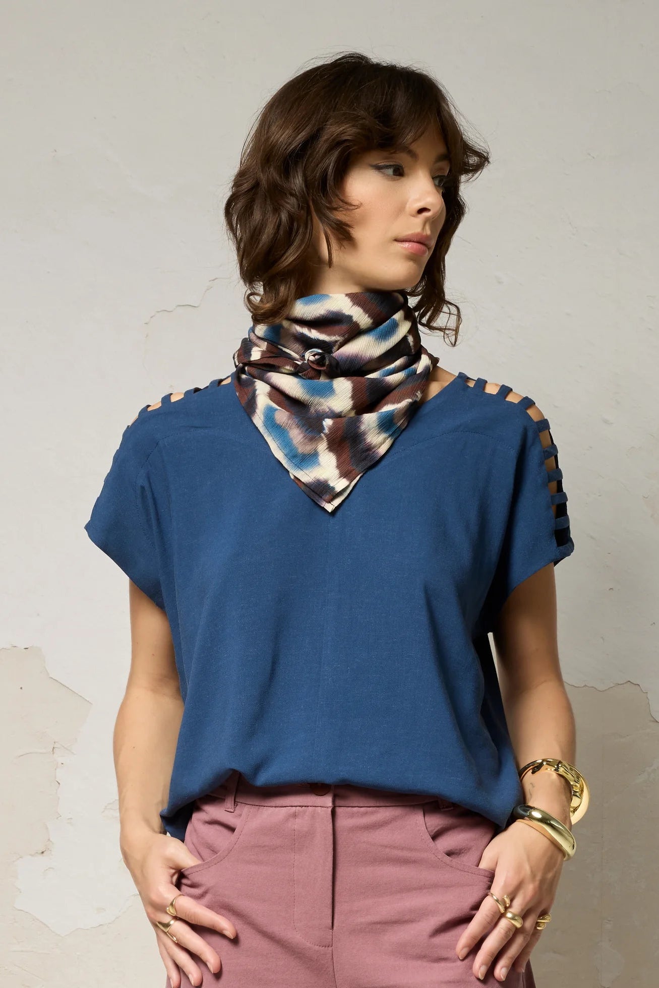 Hip-up view of a woman wearing the Mia Top by Cokluch in Denim blue, a short sleeved top with openwork ladder-like cutouts at the shoulders, a loose fit, and V-neck. She is wearing it with a patterned scarf and pink pants and standing against a plain background. 