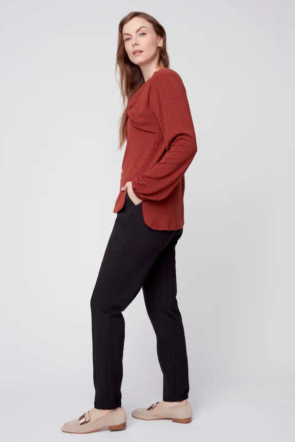 Side view of a woman wearing the Monica Sweater by Julei in Ochre, featuring a modal knit, a twist detail at the neck, bloused sleeves, and side slits. She is wearing it with black pants. and standing in front of a white background. 