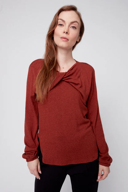 A woman wearing the Monica Sweater by Julei in Ochre, featuring a modal knit, a twist detail at the neck, bloused sleeves, and side slits. She is wearing it with black pants. and standing in front of a white background. 