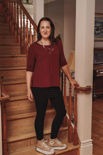 Woman wearing the Emmy Top by Moovment in Burgundy, with a boat neck, 3/4 sleeves, and side slits. She is wearing it with black pants and standing on a wooden staircase.