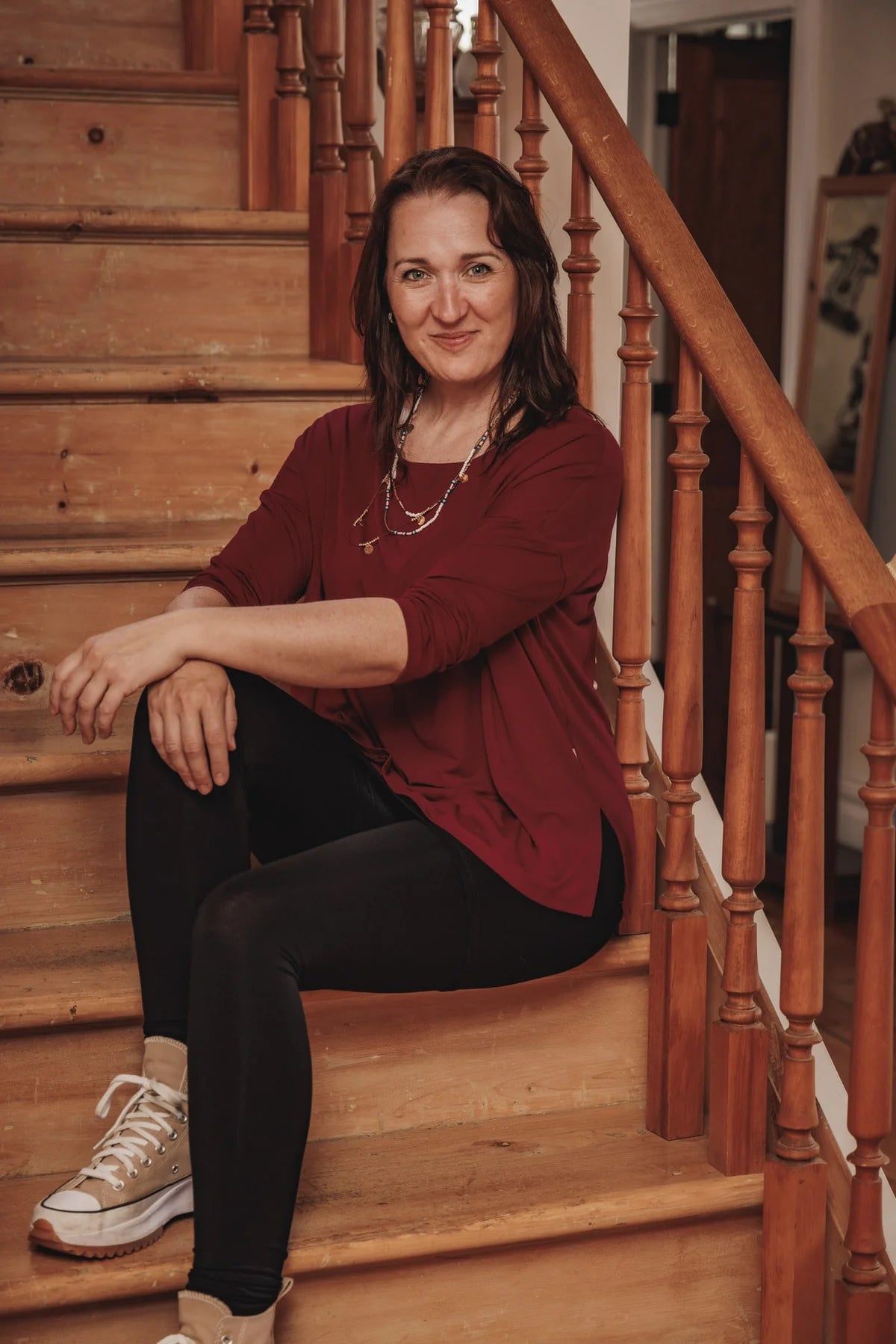 Woman wearing the Emmy Top by Moovment in Burgundy, with a boat neck, 3/4 sleeves, and side slits. She is wearing it with black pants and sitting on a wooden staircase.