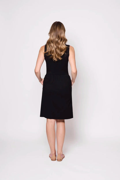 A back view of a woman wearing the Mai Dress by Tangente in Black, a  sleeveless dress with a scooped neck and gathered knee-length skirt. She is standing against a white background