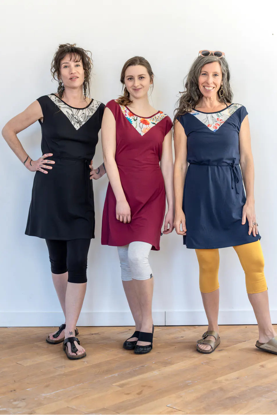 Three women wearing the Mariposa Dress by Marie C, a short sleeved short dress that's reversible back to front with printed yoke on one side, and an attached tie belt. The one in the centre is wearing it in Raspberry. 