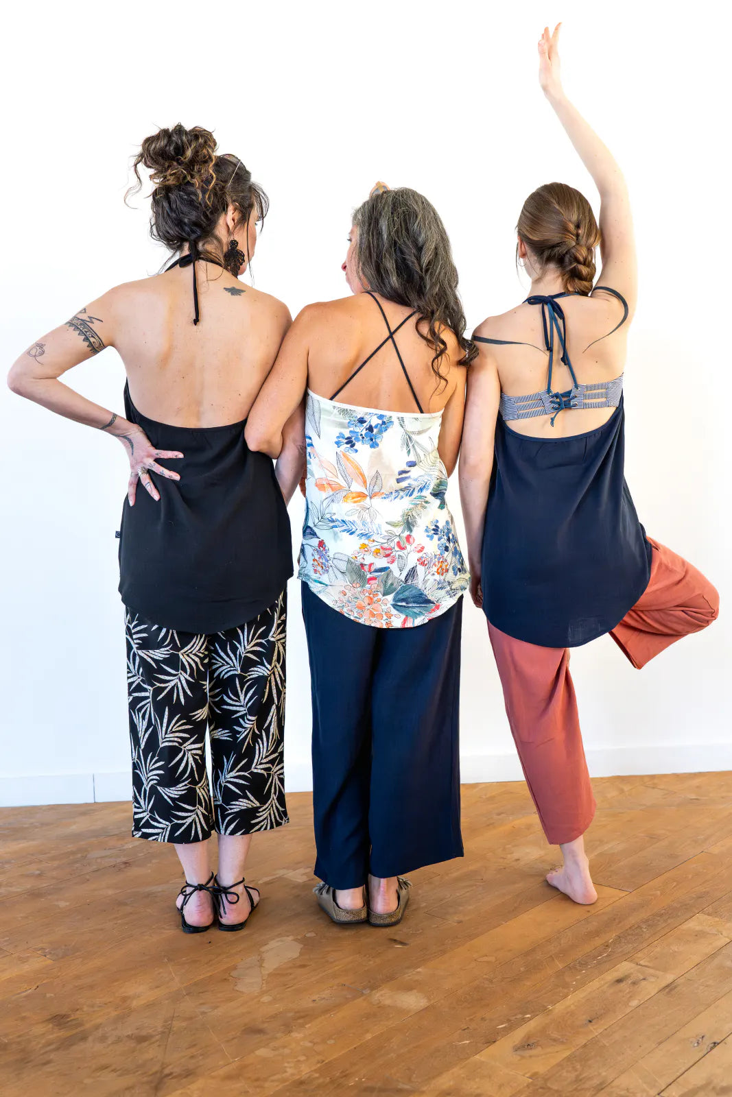 Back view of three women wearing the Verano Pants by Marie C in various colours, they are wearing them with tank tops and standing in front of a white wall. 