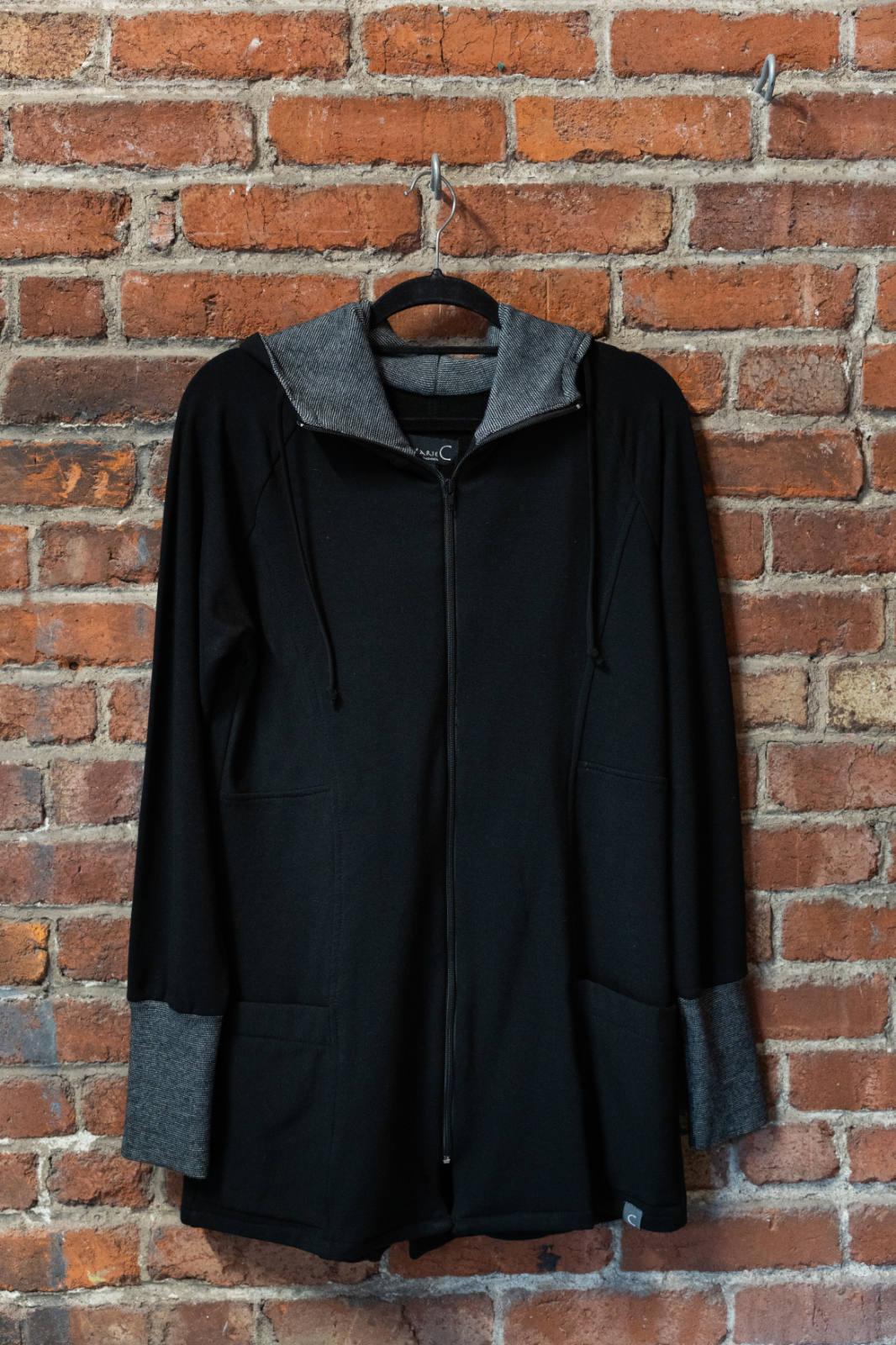 The Calma Jacket by Marie C in Black, a mid-thigh length jacket with a hood and contrasting cuffs and lining, is shown hanging against a brick wall. 