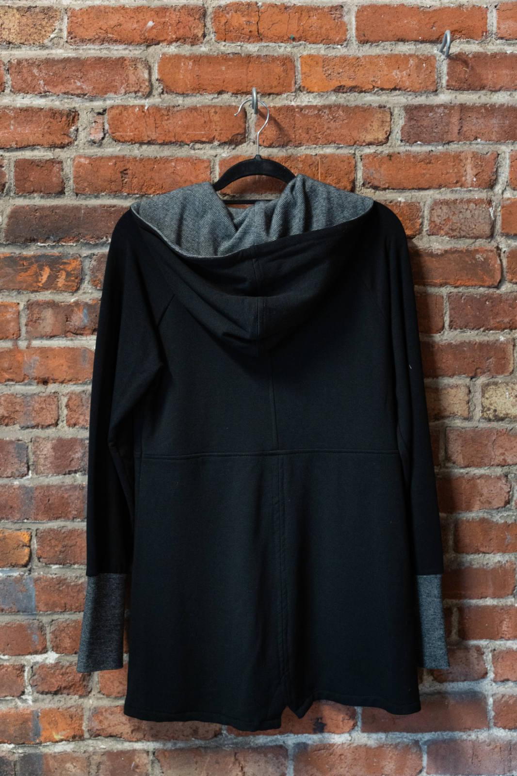 A back view of the Calma Jacket by Marie C in Black, a mid-thigh length jacket with a hood and contrasting cuffs and lining, is shown hanging against a brick wall. 