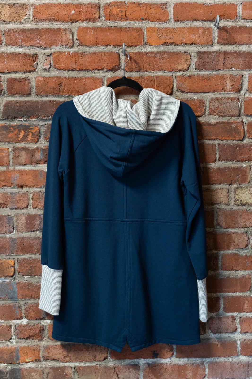 A back view of the Calma Jacket by Marie C in Navy, a mid-thigh length jacket with a hood and contrasting cuffs and lining, is shown hanging against a brick wall. 