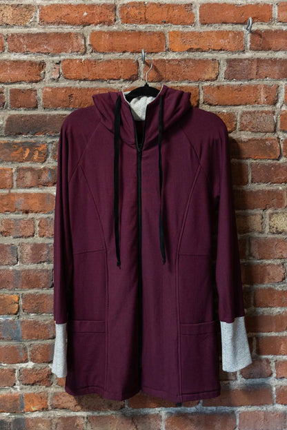 The Calma Jacket by Marie C in Wine, a mid-thigh length jacket with a hood and contrasting cuffs and lining, is shown hanging against a brick wall. 