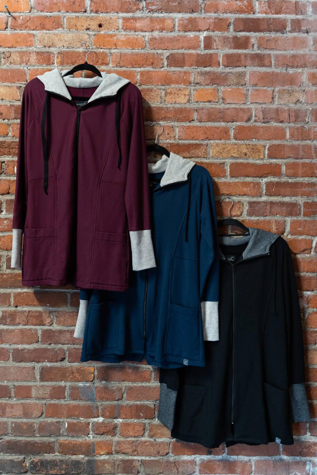 Three Calma Jackets by Marie C in Wine, Navy and Black,  a mid-thigh length jacket with a hood and contrasting cuffs and lining, are shown hanging against a brick wall. 