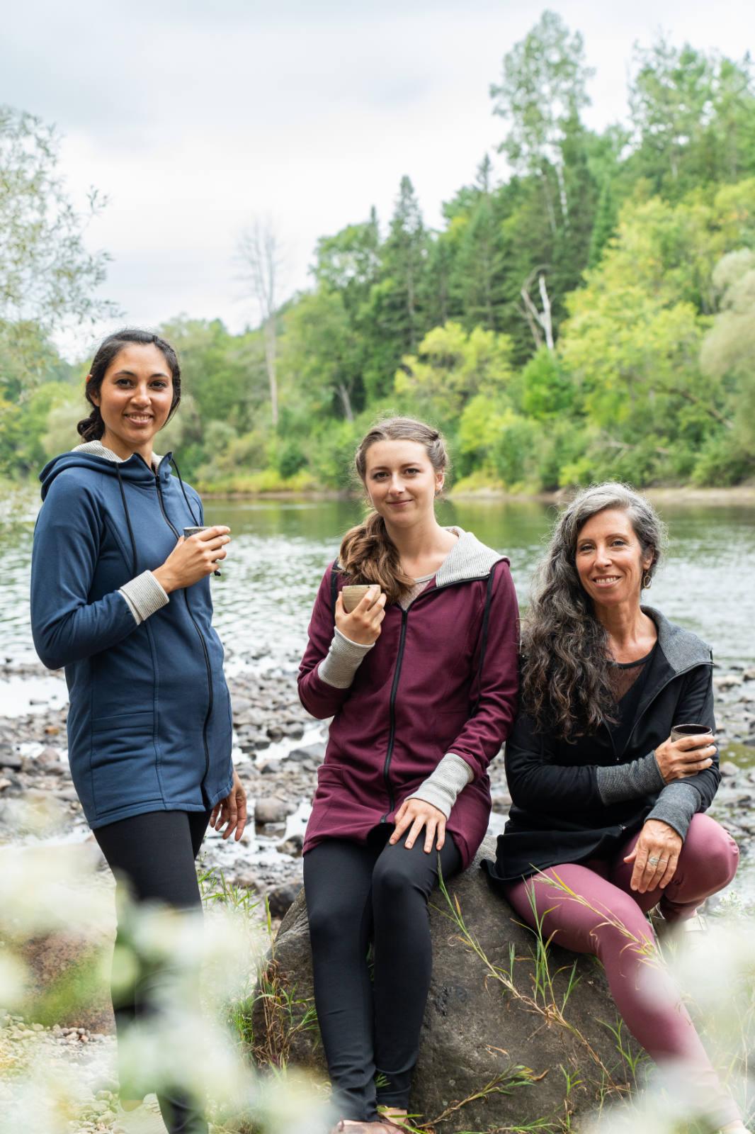 Three women wearing the Calma Jacket by Marie C in Navy, Black and Wine, a mid-thigh length jacket with a hood and contrasting cuffs and lining. They are sitting outside by a river and holding coffee cups.