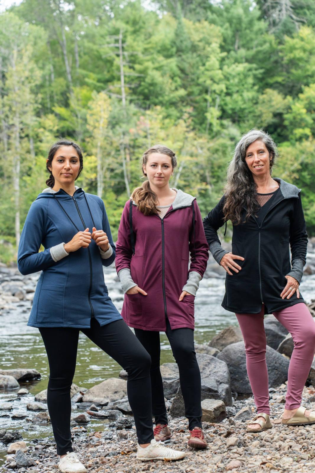 Three women wearing the Calma Jacket by Marie C in Navy, Black and Wine, a mid-thigh length jacket with a hood and contrasting cuffs and lining. They are standing outside by a river. 