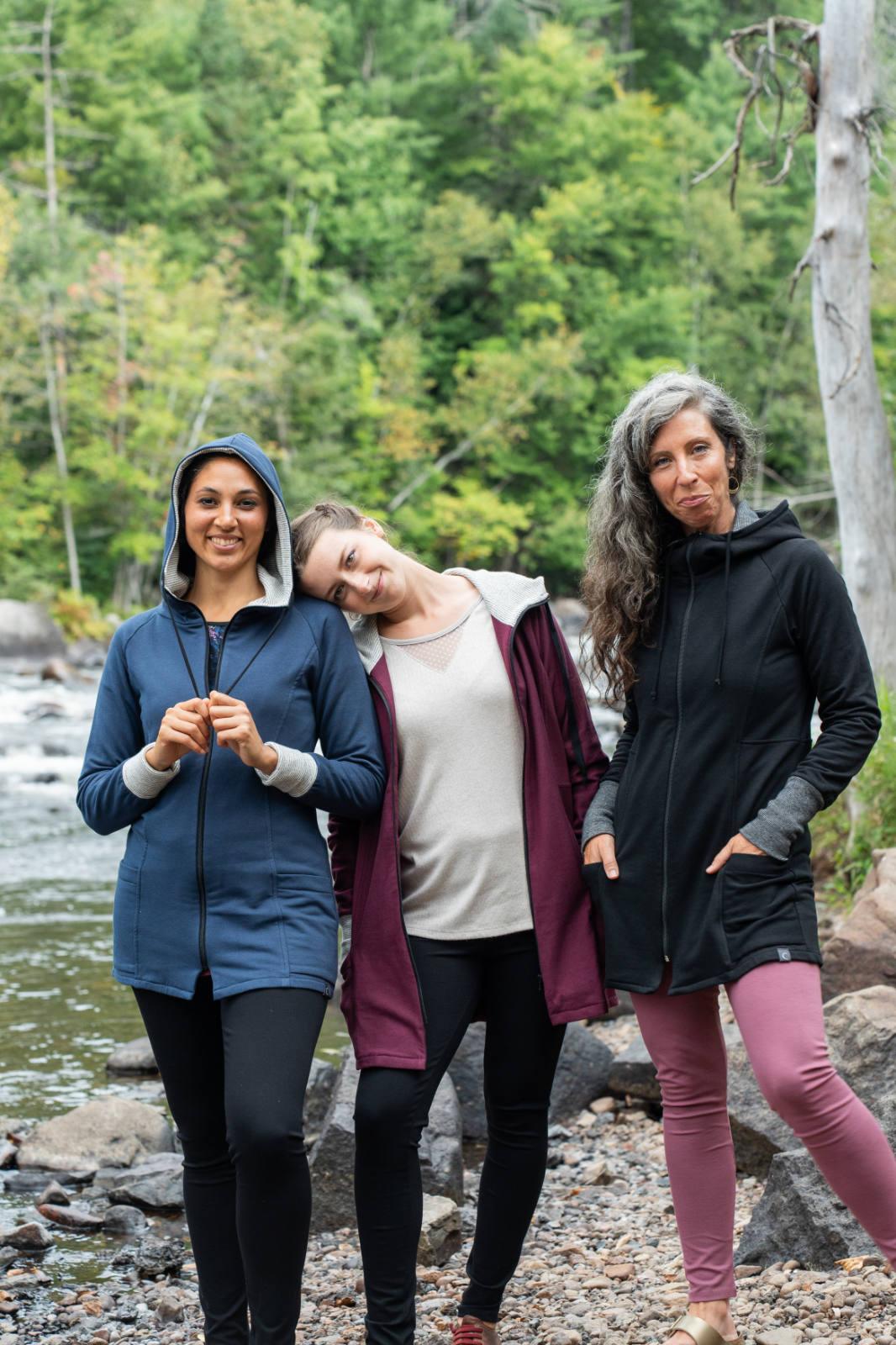 Three women wearing the Calma Jacket by Marie C in Navy, Black and Wine, a mid-thigh length jacket with a hood and contrasting cuffs and lining. They are standing outside by a river. 