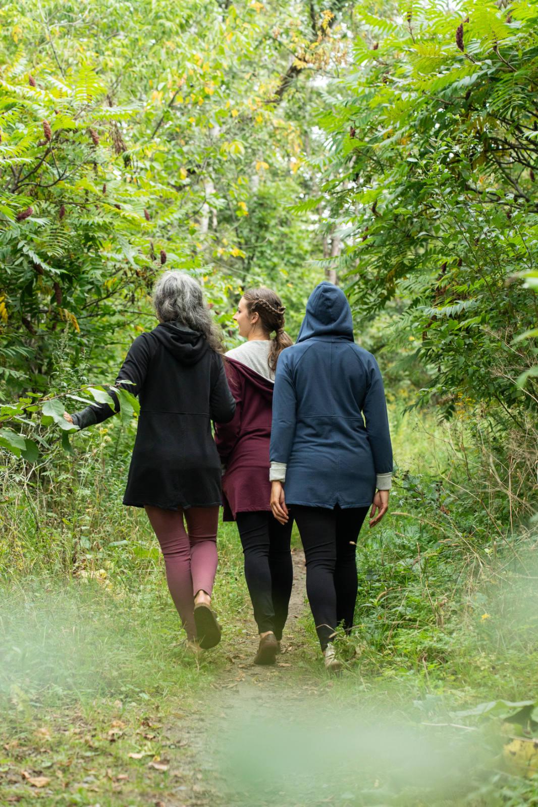 Back view of three women wearing the Calma Jacket by Marie C in Navy, Black and Wine, a mid-thigh length jacket with a hood and contrasting cuffs and lining. They are walking through a forest. 