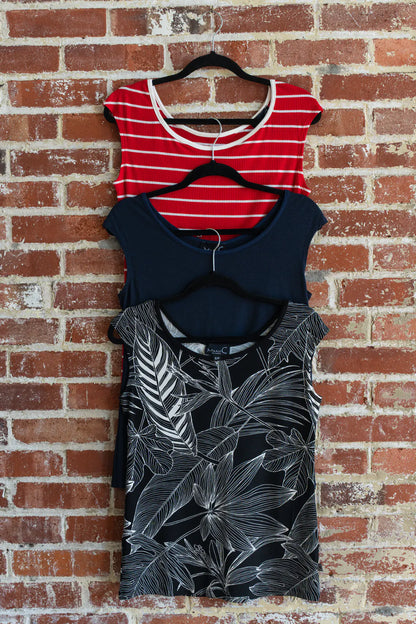 Three Sol Tops from Marie C, reversible cap sleeve tees in Red Stripe, Navy, and Black with Print,  are shown hanging vertically in front of a brick wall.