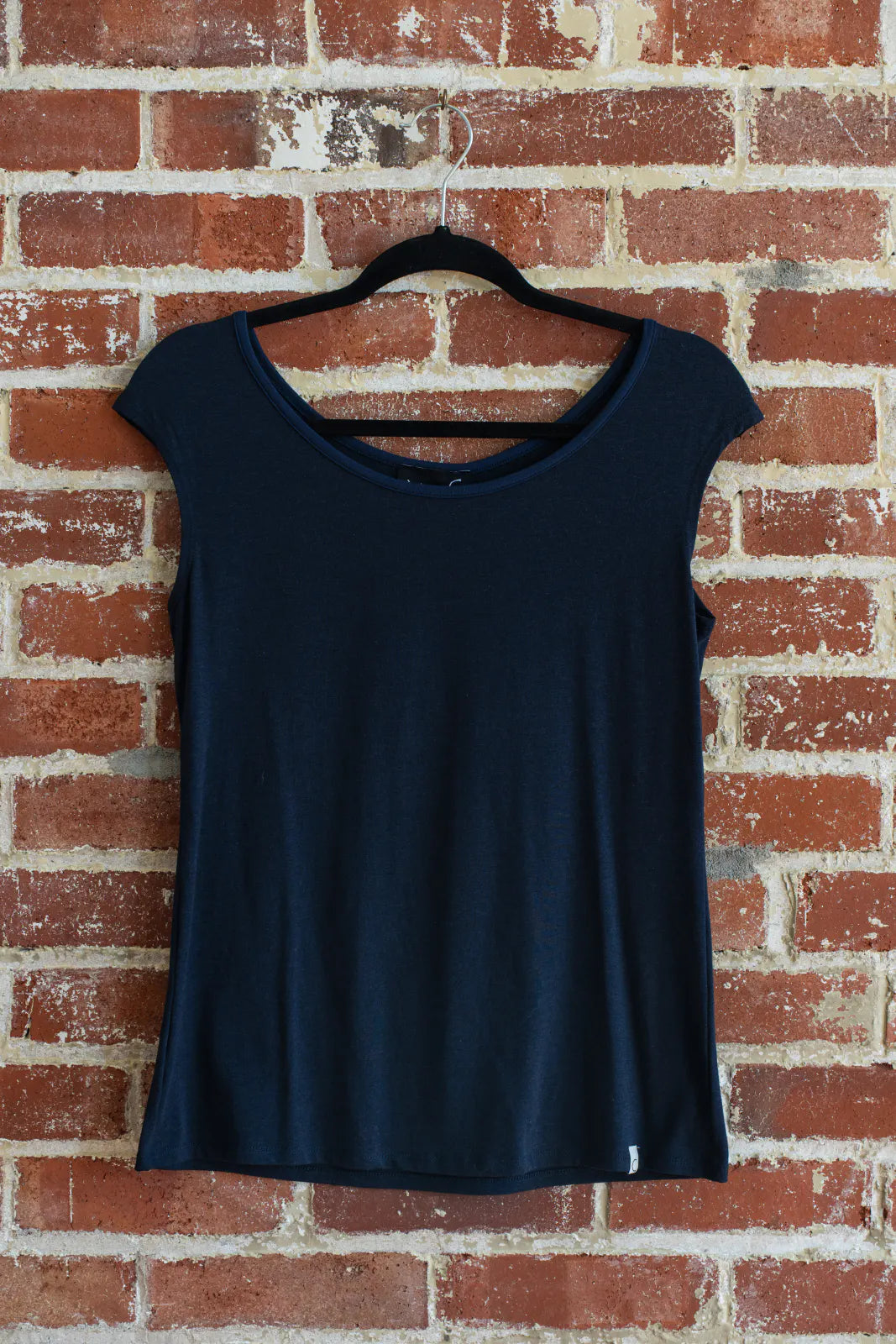 The Sol Top from Marie C in Navy, a reversible cap sleeve tee, is shown hanging vertically in front of a brick wall.