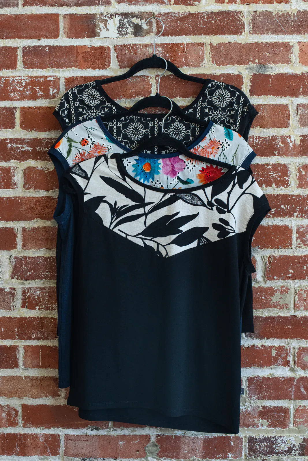 The Pepita Top from Marie C, a reversible t-shirt with a printed fabric yoke on one side, is shown hanging on a brick wall. From top to bottom the colours are: Black with Print, Navy, and Black and White. 
