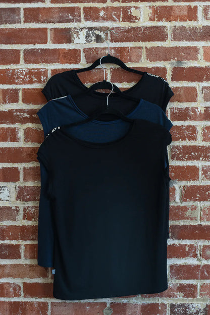 The Pepita Top from Marie C, a reversible t-shirt with a printed fabric yoke on one side, is shown hanging on a brick wall. From top to bottom the colours are: Black with Print, Navy, and Black and White. 