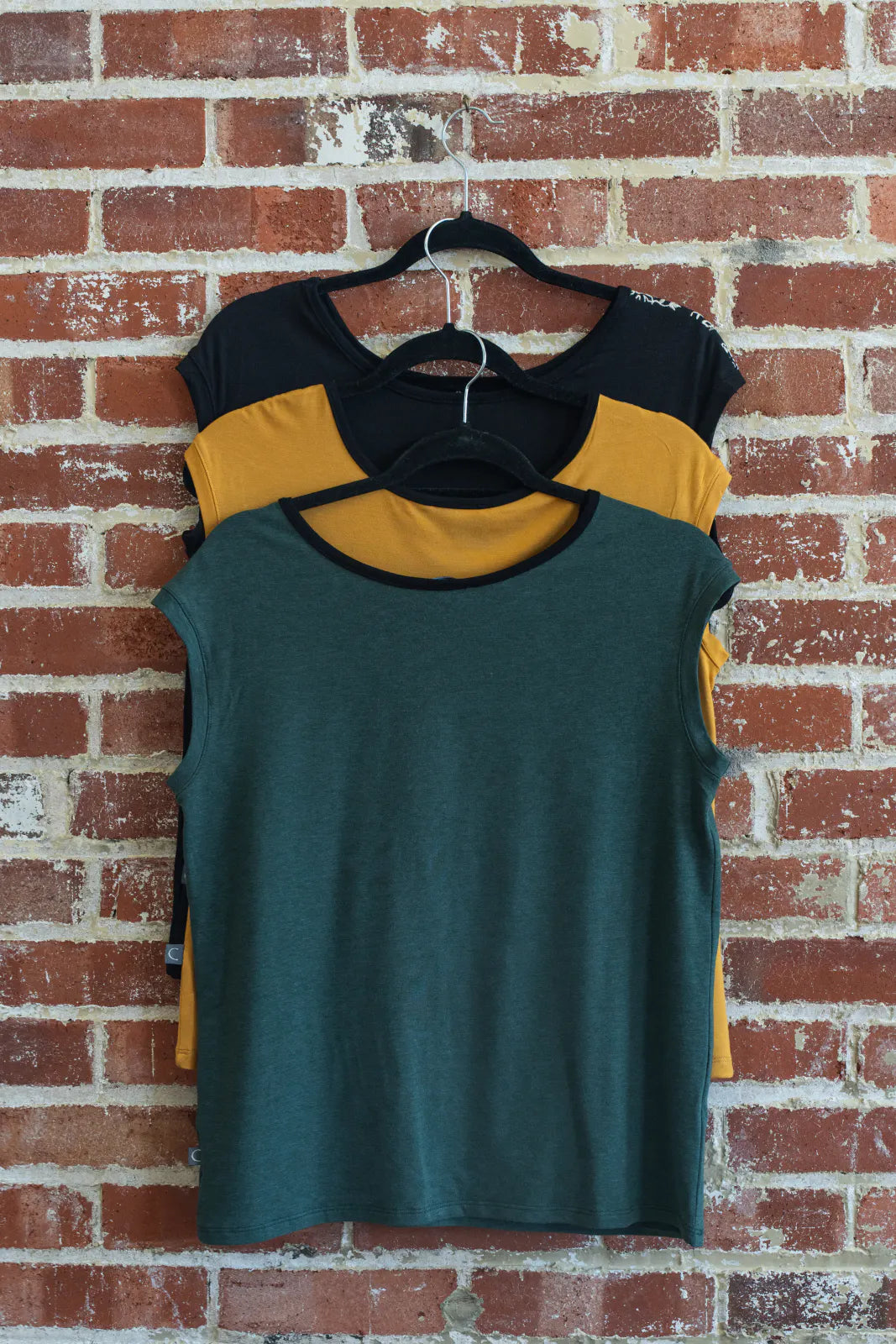 The Pepita Top from Marie C, a reversible t-shirt with a printed fabric yoke on one side, is shown hanging on a brick wall. From top to bottom the colours are: Black with Print, Yellow, Green. 