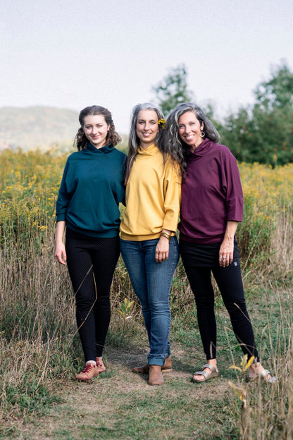 Three women wearing the BeAAA Sweater by Marie C in Everglade, Yellow and Wine, standing in a field 
