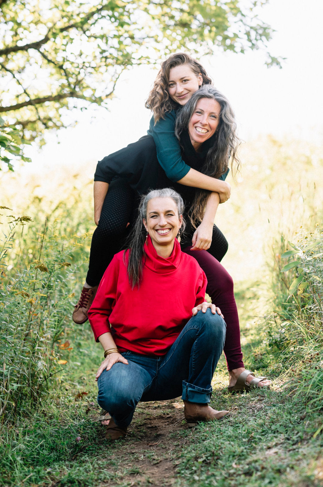Three women wearing the BeAAA Sweater by Marie C in Everglade, Black, and Red, standing in a forest.