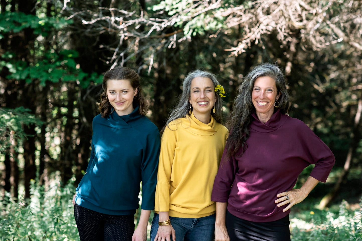 Three women wearing the BeAAA Sweater by Marie C in Everglade,Yellow, and Wine, standing in a forest. 