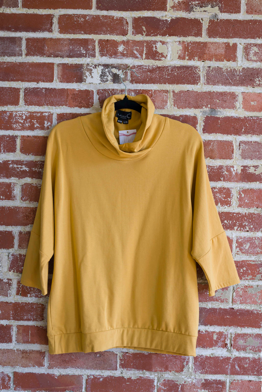 The BeAAA sweater by Marie C in Yellow on a hanger against a brick wall