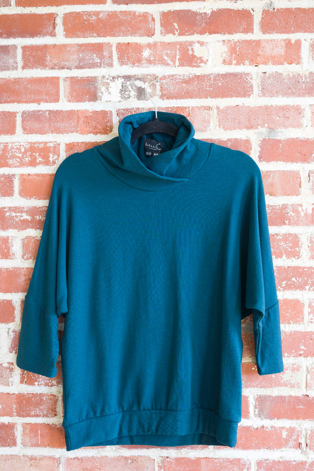 The BeAAA sweater by Marie C in Everglade on a hanger against a brick wall